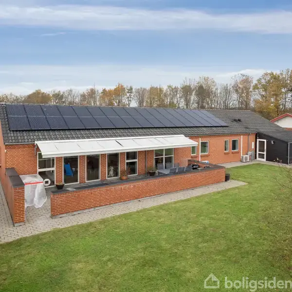 En étplans murstensvilla med solpaneler på taget står i en have. Huset har en stor terrasse med havemøbler. Haven er omgivet af hæk, og træer ses i baggrunden.