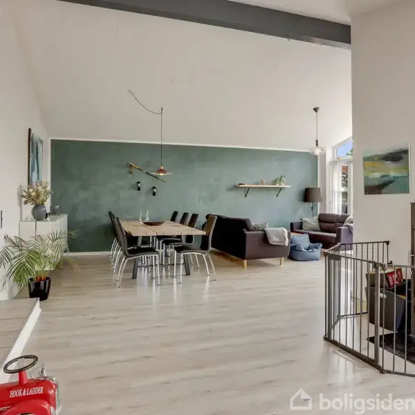 En brændeovn i stuen, omgivet af et gitter, brænder ved siden af en spisestue med et langt bord og stole samt en sofa. Lysekrone hænger fra loftet.