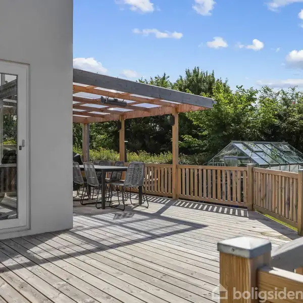 Træterrasse med spisebord og stole under en pergola i en have. I baggrunden er et drivhus omgivet af grønne træer og buske. Himlen er blå med spredte skyer.