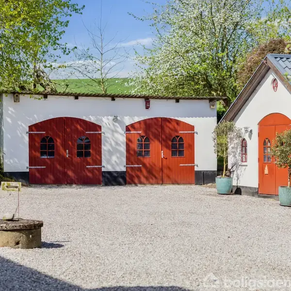 To røde garageporte står side om side på en hvid bygning omgivet af en gårdsplads med grus. Til højre er der en anden bygning med et rødt dørparti og blomsterkrukker.