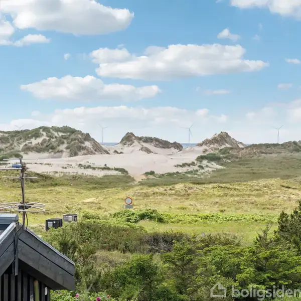 En terrasse med to liggestole og en sofa foran et sort træhus. Skiltet over døren siger "Styreboel". Baggrunden har en stenmursterrasse og vegetation under en blå himmel.