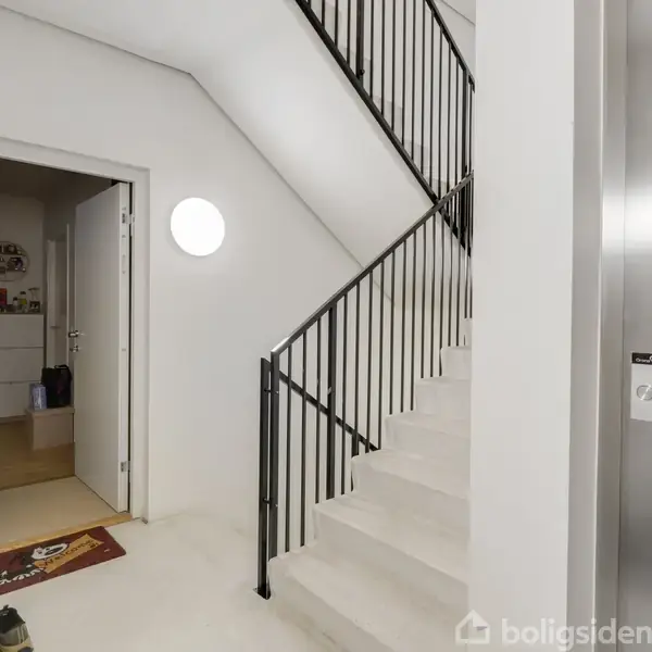 Trappeopgang med hvidmalede trin og sort gelænder. Til højre ses en elevator. I baggrunden er en åben dør til en lejlighed, hvor en skohylde og måtter er synlige.