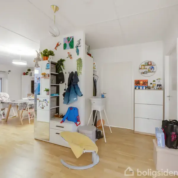 Et lyst rum med trægulv indeholder en åben stue, reoler med tøj, og spisebord i baggrunden. Væggene er dekoreret med planter og kunst. Tekst på billedet: "boligsiden".