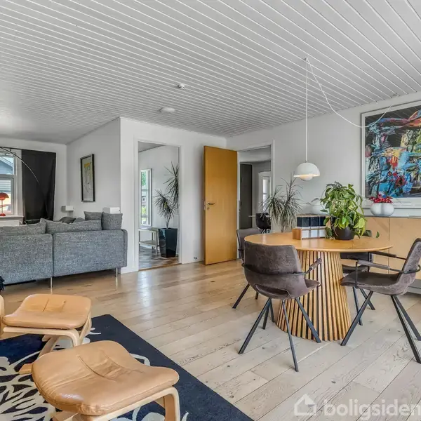 Stue med to lænestole og skammel i forgrunden, grå sofa i baggrunden. Træbord med stole og en plante. Kunstnerisk billede hænger ved skænk, modelskib ovenpå. Trægulv og træloft.