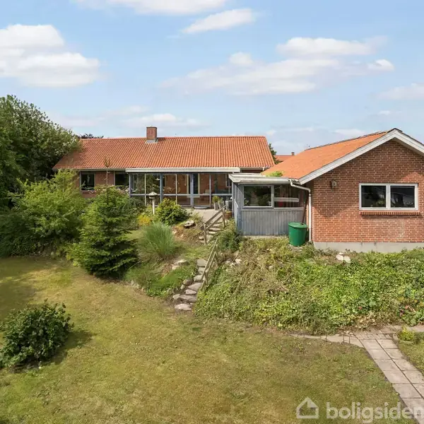 Rødt hus med skråt tag står i en grøn have. Stensti fører op til indgangen. Omkringliggende buske og træer skaber en hyggelig atmosfære under en blå himmel med lette skyer.