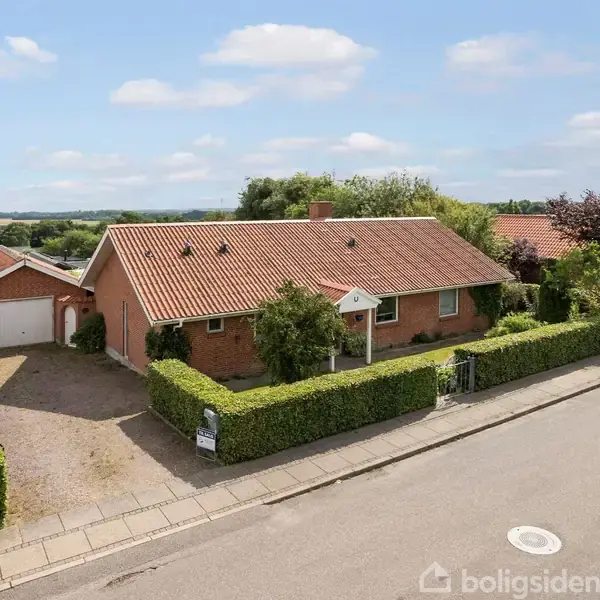 Et rødt murstenshus med tegltag står blandt grønne hække. En garage er ved siden af huset. Beliggende i et villakvarter med udsigt til åbne marker og himmel.