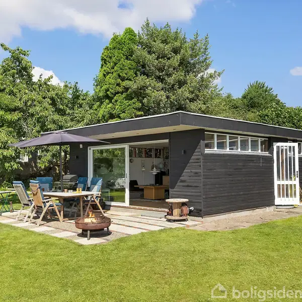 Sort sommerhus står stille omgivet af en grøn have med træer. Foran huset står et bord og stole under en parasol, og der er også en bålfad på terrassen.