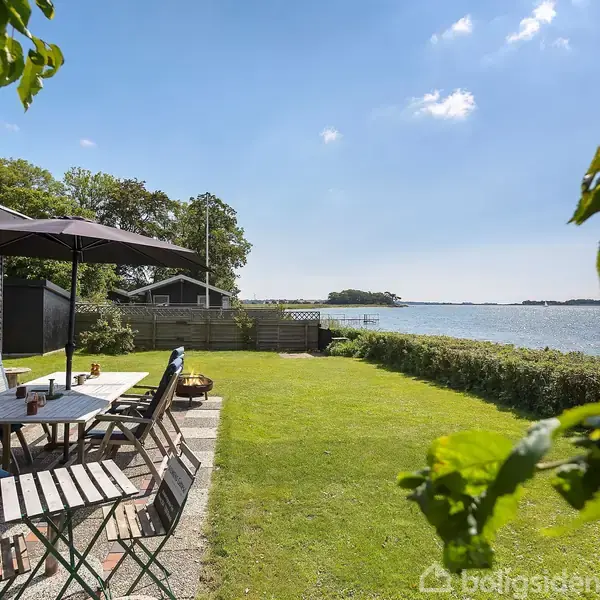 Et bord med stole står på en terrasse under en parasol, ved siden af en græsplæne. Omgivelserne viser en udsigt over en sø med blåt vand og en klar himmel.