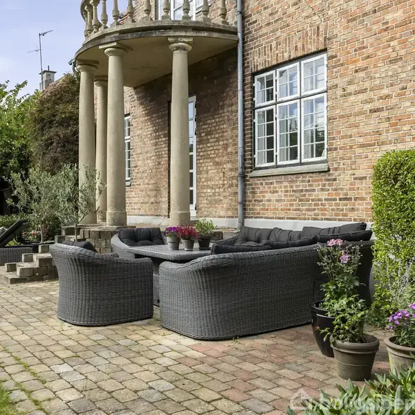Rattanlignende havemøbler står placeret i en u-form på en terrasse med brostensbelægning ved en muret bygning. Der er planter i potter og tætte buske i baggrunden.