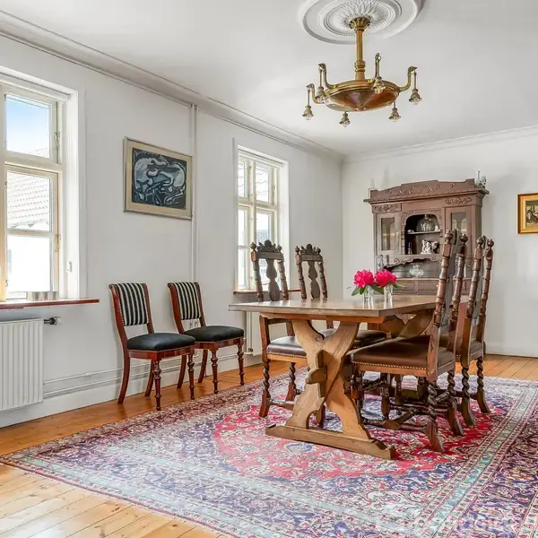 Et spisebord med træstole står på et orientalsk tæppe i et lyst rum. Der er et vitrineskab, en kakkelovn og malerier på væggene, med store vinduer.