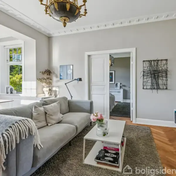 En grå sofa med tæppe står i en lys stue. Et moderne sofabord med bøger og blomster foran, og et vindue til venstre giver udsigt til en have.