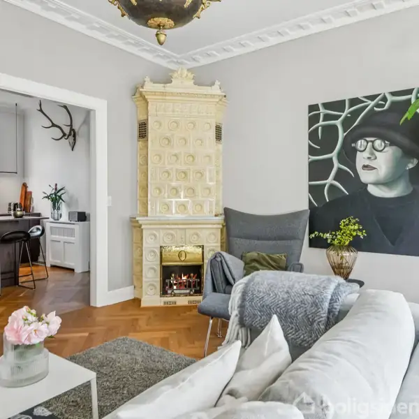 En stilfuld stue med en stor sofa, flisebelagt kakkelovn, og et maleri på væggen. Et stort, grønt træ står ved siden af, og køkkenet ses i baggrunden.