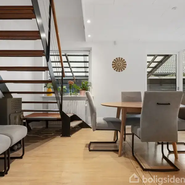 En moderne stue med trappe i træ og metal til venstre. Grå stole omgiver et rundt træbord. Vinduer med udsigt til et tag og en vægdekoration ses bagerst.