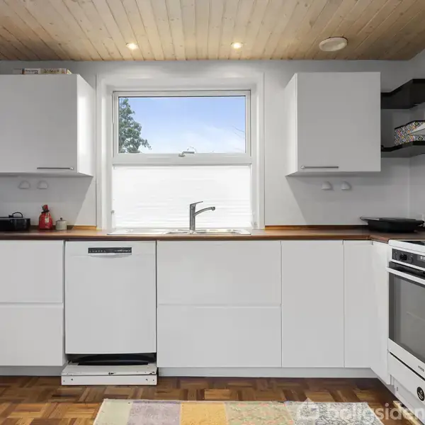 Køkken med hvide skabe, opvaskemaskine og komfur, med træbordplade og vindue der viser blå himmel. Trægulv og trælister i loftet skaber en lys og åben atmosfære.