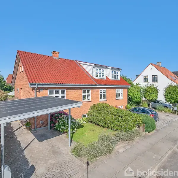 Rødt murstenshus med rødt tag står på en pæn gade, omgivet af velplejede buske. Carport ved siden af huset rummer en bil. Blå himmel og flere huse i baggrunden.