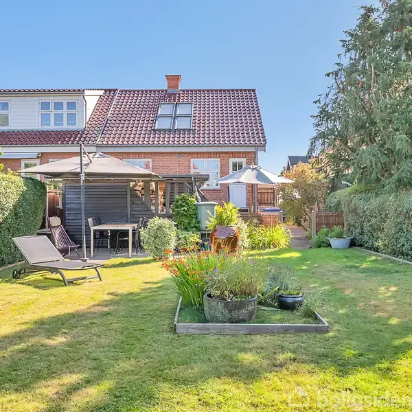 Hus med rød murstensfacade og rødt tegltag. I haven er der en græsplæne, hækkesektioner, en pavillon med havemøbler og solseng. Bagved ses blå himmel og træer.