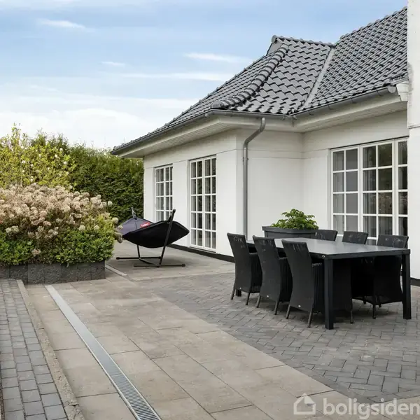 Hus med sort tegltag og hvid facade, til højre et udendørs bord med stole på en flisebelagt terrasse, omgivet af en veltrimmet grøn hæk og blomsterbede.