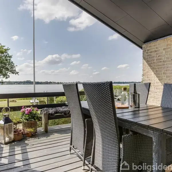 Bord med flettede stole står på en terrasse under et halvtag. I baggrunden ses en græsplæne og en sø med træer under en delvis skyet himmel.