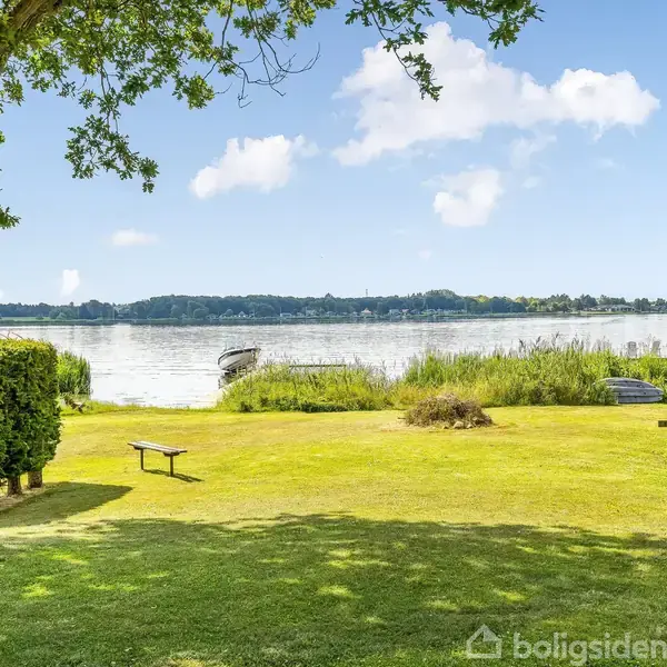 Græsplæne med picnicbord i træ, står stille nær en sø omgivet af buske og træer i en rolig, solrig natur. En båd ligger ved kysten, mens himlen er klar med skyer.