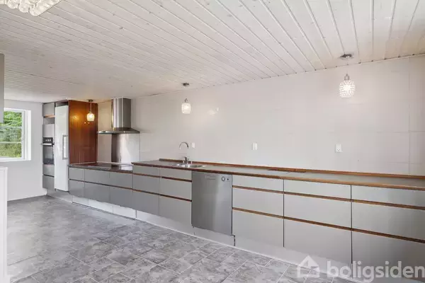 Et moderne køkken med grå skabe og en opvaskemaskine. Køkkenet har en emhætte, hvide vægge og loft, samt grå flisegulv. Store vinduer lyser rummet op i baggrunden.