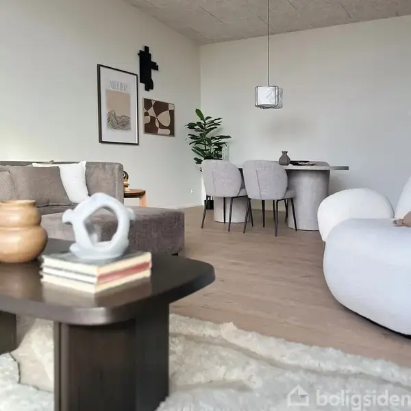 En moderne stue med sofa, sofabord med keramikdekorationer og bøger. Et rundt spisebord med stole står i baggrunden, en plante og vægkunst dekorerer rummet.