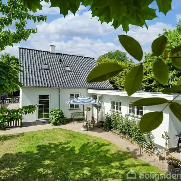 Hus med hvid facade og sort tegltag, udenfor står et parasol over et bord i en solrig have omkranset af grønne træer og buske.