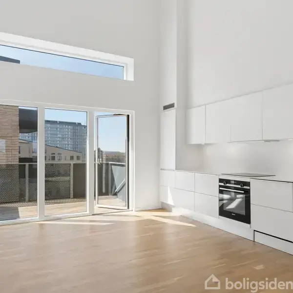 Et moderne køkken med hvide skabe og indbygget ovn er placeret til højre, mens glasdøre åbner ud til en balkon. Rummet har lyst trægulv og en høj loftshøjde.