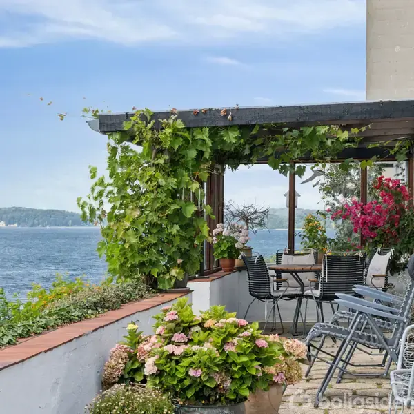 Terrasse med borde og stole under en pergola med klatreplanter, beliggende ved havet. I baggrunden ses en vandudsigt med skovklædte kystlinjer og sejlbåde på vandet.