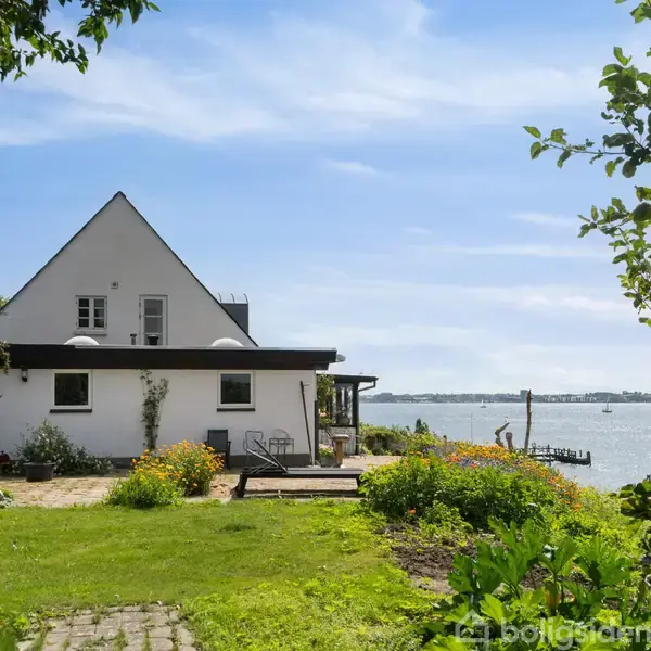 Et hvidt hus med trekantet tag står ved en frodig have og terrasse. Huset ligger ved vandet, med udsigt til en mole og blå himmel.