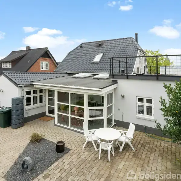 Et hvidt hus med tagterrasse har en glasudbygning foran. Foran huset står et rundt hvidt bord med fire stole på en flisebelagt terrasse omgivet af grønne hække.