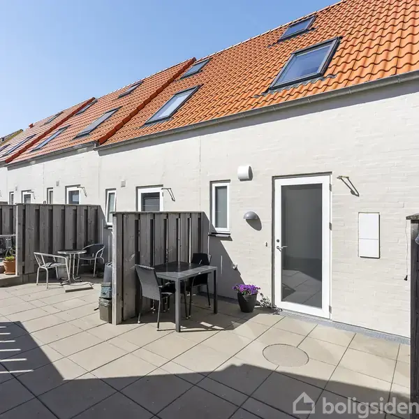 Rækkehus med rødtag, verandamøbler står på flisebelagt terrasse. Flere små vinduer indrammer hvide væggen. Sollys skaber skygger på jorden.