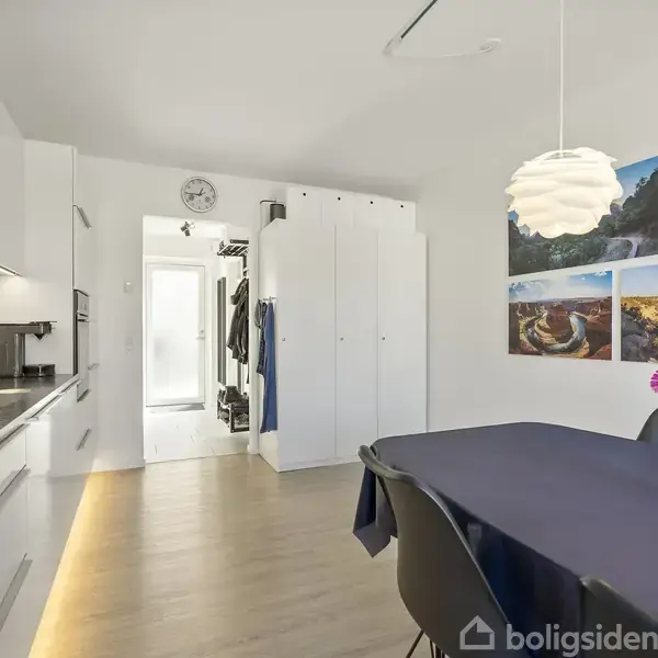 Et moderne køkken med hvide skabe og en bordplade ved siden af et spisebord dækket med en blå dug. Baggrunden rummer farverige blomster og naturfotografier på væggen.