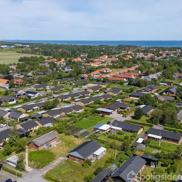 Boligområde strækker sig mod horisonten med huse arrangeret langs veje, omgivet af grønne områder. I baggrunden ses skov og hav, der giver et naturskønt perspektiv.