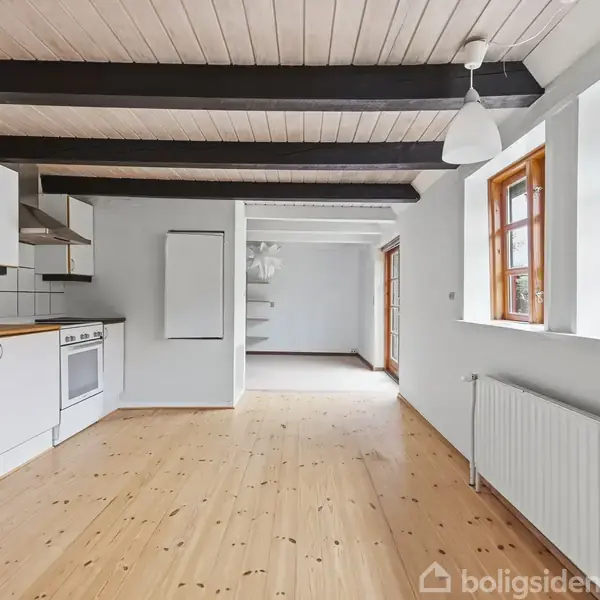 Køkken med hvide skabe og ovn langs en væg. Trægulve i lyse planker, sorte loftsbjælker. Store vinduer med trærammer, der giver lys fra haven udenfor.