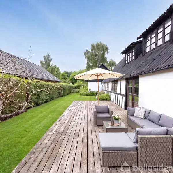 En terrasse med grå loungemøbler og parasol. Den er placeret ved et moderne sort-hvidt hus, omkranset af en velplejet græsplæne og hæk. Tekst: "Visualisering".