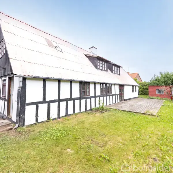Bindingsværkshus med hvid facade og sorte træstrukturer står stille på en grøn græsplæne omgivet af træer og buske. Til højre ses et rødmalet skur. Ingen tekst ses.