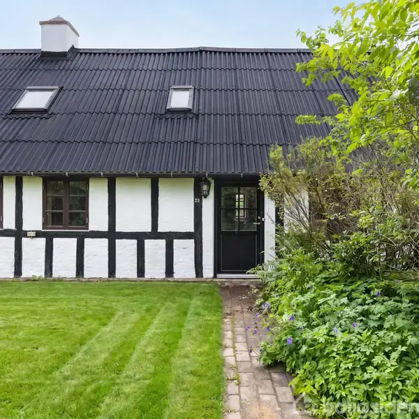 Sort-hvid bindingsværkshus med sorte tagsten. Dør og vinduer på facaden. Beliggende i en grøn have med græsplæne og buske. Tekst: "Visualisering".