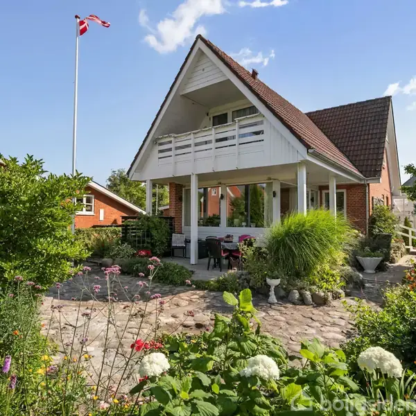 Et hvidt hus med trekantet tag, terrasse og balkon står i en frodig have fyldt med blomster og buske. I baggrunden vajer et dansk flag på en høj flagstang under en blå himmel.