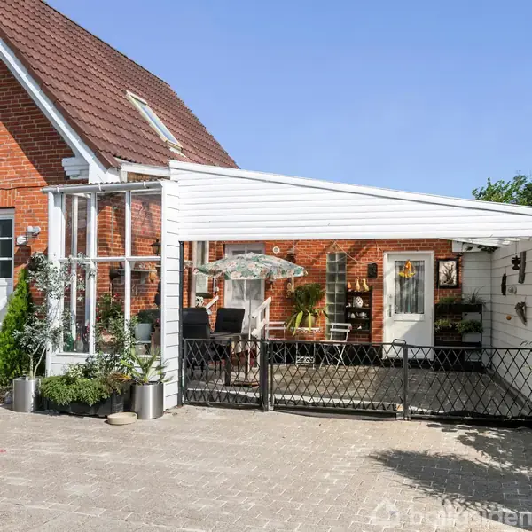 En murstensbygning med en carport af træ står foran et hus med rødt tag. Der er planter i krukker ved indgangen, og en gårdhave ses til højre.
