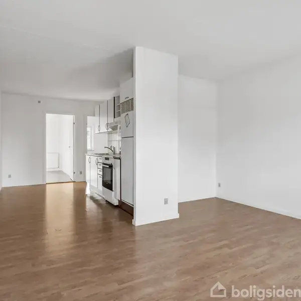 Et åbent rum med trægulv indeholder et hjørnekøkken med ovn og skabe. Udsigten strækker sig mod en lys gang, med hvide vægge og en radiator til venstre.