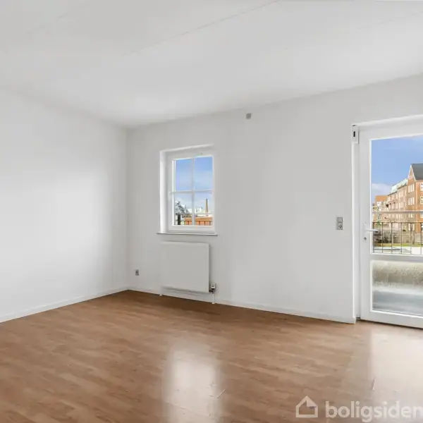 Et tomt værelse med trægulv, hvidmalede vægge og vindue til venstre. En glasdør til højre fører ud til en altan med udsigt over bygninger udenfor.