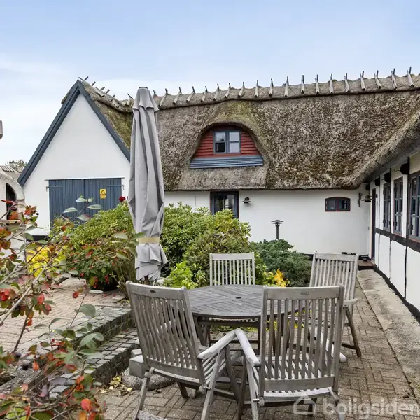 Et rundt havebord med træstole står på en flisebelagt terrasse foran et bindingsværkshus med stråtag. Omkring terrassen er der blomsterbede og en udendørs grillpejs.