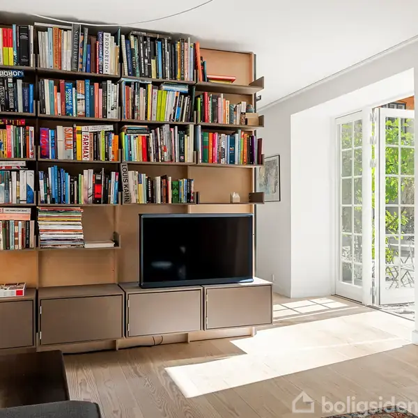 Bogreol fyldt med bøger hænger på en hvid væg, med et tv nedenunder. Rummet er lyst med trægulv, og en åben dør fører til en udendørs terrasse med grønne omgivelser.