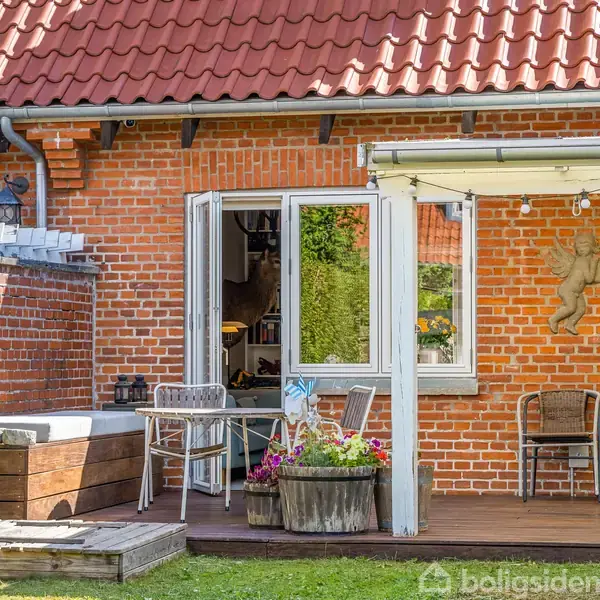 Hus med rød teglsten og tag, åbne franske døre fører til en terrasse med stole og blomsterkasser. På væggen hænger en dekorativ figur. Græsplæne omgiver terrassen.