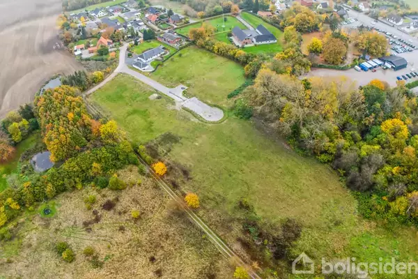 Grønne marker strækker sig i et åbent område omgivet af træer i efterårsfarver. I baggrunden ses en lille landsby med huse og parkeringsplads. Ingen tekst fundet i billedet.