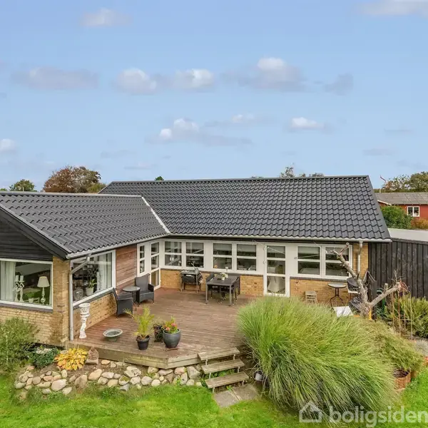 Et moderne parcelhus med sort tegltag står stille i en have med terrasse. Omkring huset er der grøn græsplæne, buske og blomster, der skaber en hyggelig atmosfære.