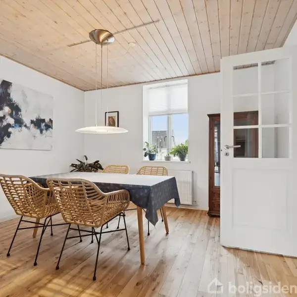 Et spisebord med fire flettede stole står i et lyst rum med træloft. En lampe hænger over bordet, og et abstrakt maleri pryder væggen. En dør står åben.