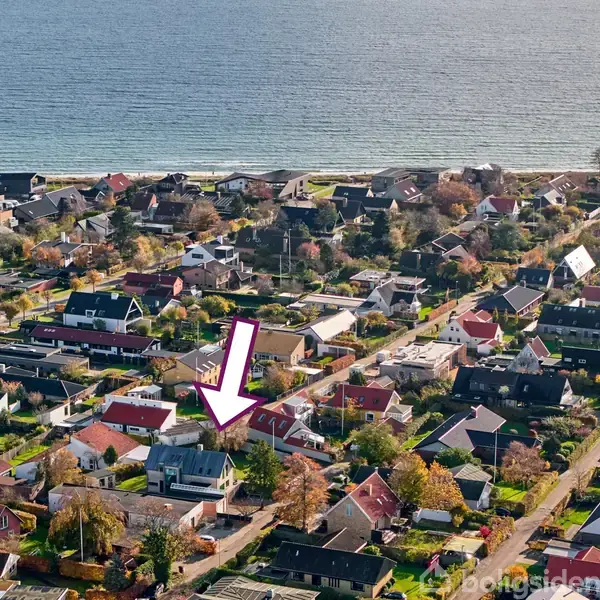 Hus fremhævet med en stor, lyserød pil befinder sig i et kystnært boligområde med mange boliger, tæt på vandet. Umgebung domineres af huse og veje under en klar himmel.