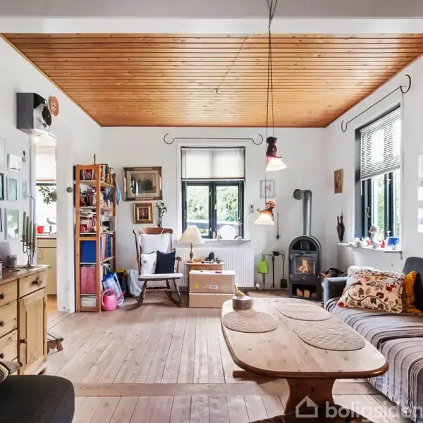 Stue med træloft, sofa og sofabord i midten. Bøger og dekorationer på hylderne. Brændeovn står i hjørnet ved et vindue, skaber en hyggelig atmosfære.