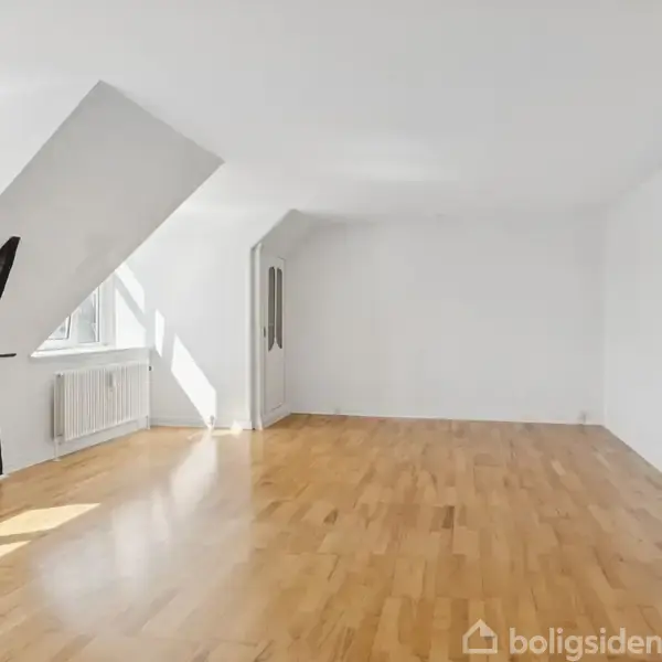 Et stort, lyst værelse med trægulv og skråvægge. To vinduer lader sollys komme ind, og en sort, skulpturel bjelke står ved siden af dem. Væggene er hvide.